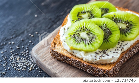 whole-grain toast with cashew cream and kiwi slices, dusted with chia seeds on a rustic wooden board, dark textured background, deep moody lighting, scattered seeds, concept of healthy breakfast 123734740