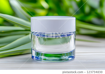 Clear cosmetic jar surrounded by green leaves, ideal for organic skin serum and therapy products, promoting a fresh spa experience with essential oils 123734830
