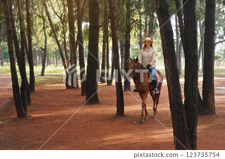 Young woman in shirt and straw hat, rides brown horse in the morning park, blurred trees in background 123735754