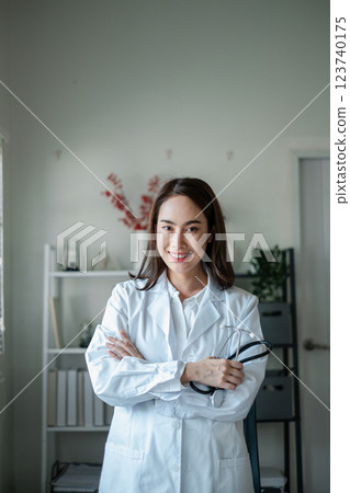 Smiling female doctor in a white coat holding a stethoscope, standing in a modern medical office with a bookshelf and plants. Smiling female doctor in a white coat holding a stethoscope, standing in a modern medical office with a bookshelf and plants. 123740175