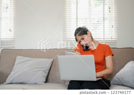 A young woman in an orange shirt multitasks with a laptop and phone on a cozy sofa in a bright living room. A young woman in an orange shirt multitasks with a laptop and phone on a cozy sofa in a bright living room. 123740177