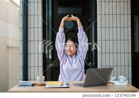 Woman in casual attire stretches at a desk with a laptop and coffee, promoting relaxation and productivity in a modern home office. Woman in casual attire stretches at a desk with a laptop and coffee, promoting relaxation and productivity in a modern home office. 123740194