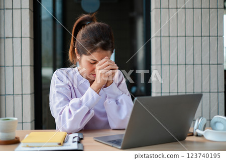 A young woman sits at a desk with a laptop, appearing stressed and deep in thought, in a modern office environment. 123740195