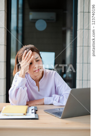 A woman in a light purple blouse looks stressed while working on a laptop at her office desk. 123740196