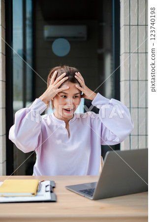 A woman in a light purple blouse looks surprised while using a laptop at an outdoor cafe table, with documents and a notebook nearby. A woman in a light purple blouse looks surprised while using a laptop at an outdoor cafe table, with documents and a notebook nearby. 123740198