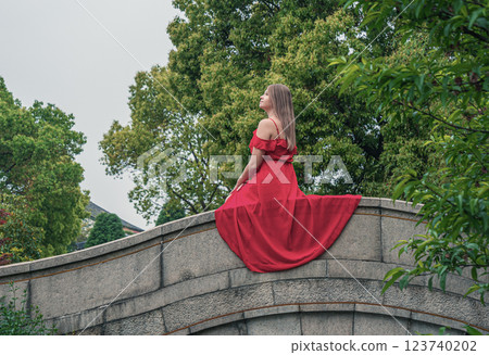Woman in a flowing red dress on a stone bridge in a tranquil garden during cloudy weather 123740202