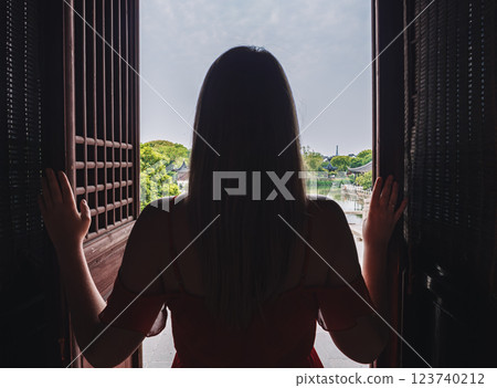 Young woman standing at the doorway overlooking a tranquil landscape with water and greenery during daylight hours 123740212