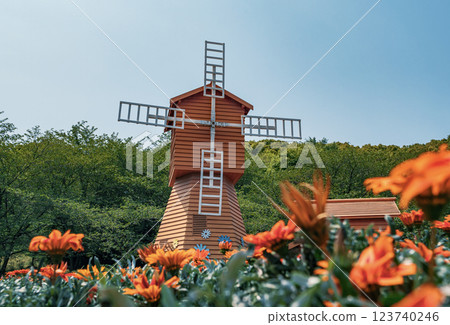 Bright orange flowers surround a wooden windmill against a clear blue sky in a lush greenery setting 123740246