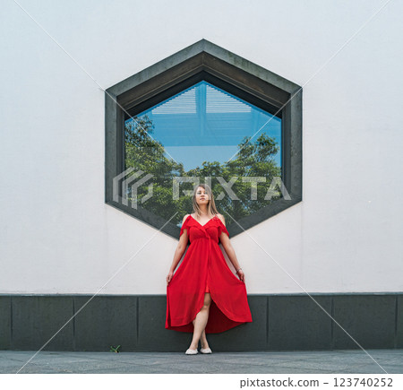 A woman stands elegantly in a flowing red dress, striking a pose in front of a unique hexagonal window. A woman stands elegantly in a flowing red dress, striking a pose in front of a unique hexagonal window. 123740252