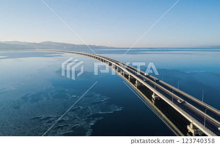 Aerial view of a long road bridge stretching over calm waters at dawn with distant mountains and city skyline 123740358