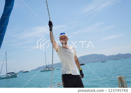 Male captain on deck of sailboat opening sails pulling the rope Male captain on deck of sailboat opening sails pulling the rope 123745748