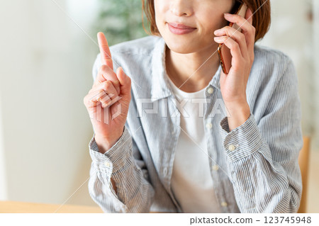 一位中年婦女在家中的客廳微笑著用食指指著，一邊用智慧型手機通話 123745948