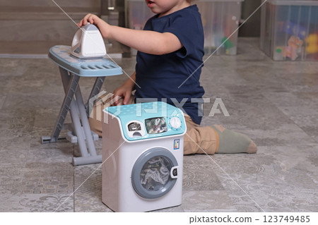 Little boy plays with toy iron and washing machine at home on floor. Cleaning, household chores, kids and educational games. Little boy plays with toy iron and washing machine at home on floor. Cleaning, household chores, kids and educational games. 123749485