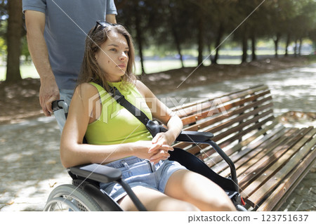 Nurse pushing young woman in wheelchair in park near bench 123751637