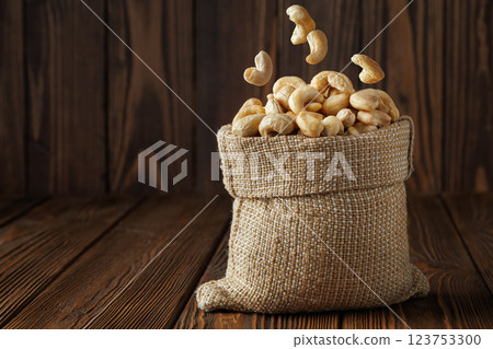 Cashew nuts are falling into burlap sack on rustic wooden table, creating visually appealing arrangement against a dark background 123753300