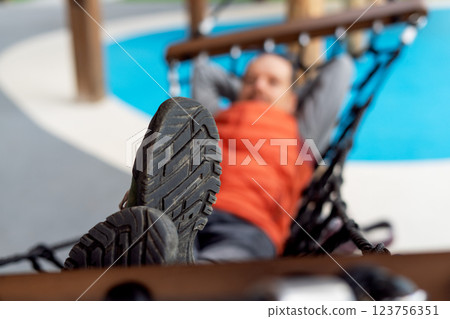 Close-up of rugged hiking boot sole in focus, with blurred man relaxing in hammock at an outdoor playground, concept for relaxation, adventure, and outdoor recreation themes Close-up of rugged hiking boot sole in focus, with blurred man relaxing in hammock at an outdoor playground, concept for relaxation, adventure, and outdoor recreation themes 123756351