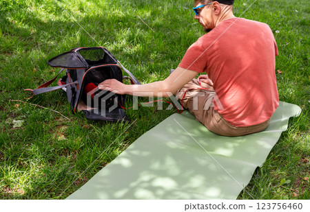man in red T-shirt sits on green yoga mat in park, accompanied by black cat inside pet carrier on green grass, cat wearing red harness, peeks out curiously while man gently pets it , pet companionship man in red T-shirt sits on green yoga mat in park, accompanied by black cat inside pet carrier on green grass, cat wearing red harness, peeks out curiously while man gently pets it , pet companionship 123756460