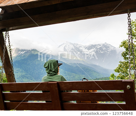 woman in green hoodie with backpack sitting on wooden swing and enjoying the mountain scenery in mountain hike copy space 123756648