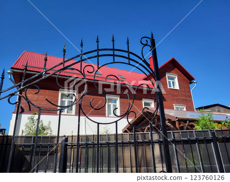 Wrought-iron gates on the background of a brown house with a red roof against the blue sky. 123760126