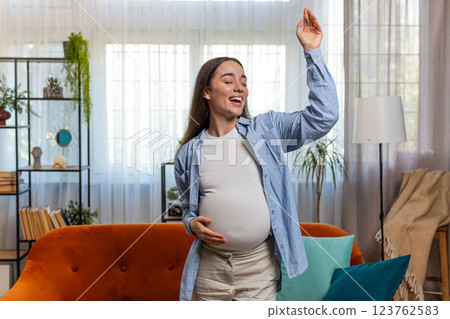 Happy smiling young pregnant woman playfully dancing listening music touching her big belly at home Happy smiling young pregnant woman playfully dancing listening music touching her big belly at home 123762583