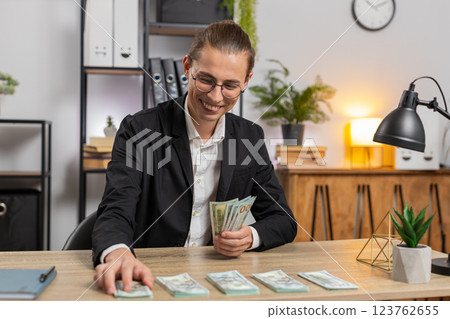 Successful young businessman guy in formal suit sitting counting money at table in home office Successful young businessman guy in formal suit sitting counting money at table in home office 123762655
