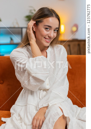 Happy attractive young woman sitting on sofa couch in living room looking shy at camera and smiling 123762775