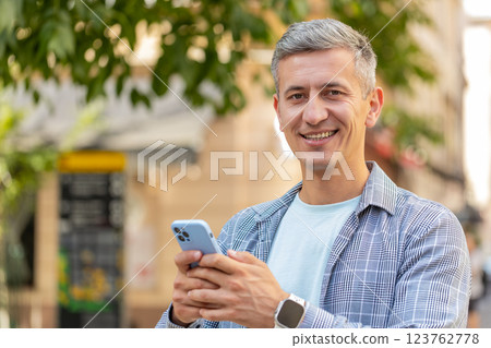 Smiling mature man using smartphone browsing internet, finishing work, looking at camera outdoors 123762778
