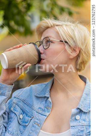 Happy Caucasian young blonde woman enjoying morning coffee hot drink and walking on city street 123762795