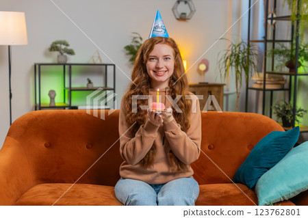 Happy woman wears birthday hat makes wish congratulating blowing burning candle on cake at home Happy woman wears birthday hat makes wish congratulating blowing burning candle on cake at home 123762801