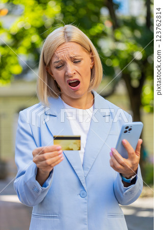 Frustrated mature businesswoman in suit using credit card and mobile phone standing on city street 123762814