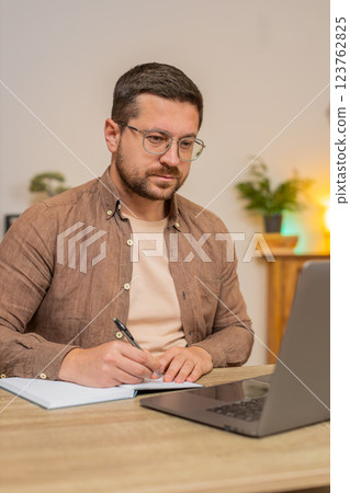 Young freelancer writing down notes while attending online meeting on laptop at home office desk 123762825