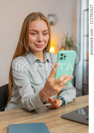 Happy female freelancer sitting at desk using smartphone texting smiling in living room at home 123762827