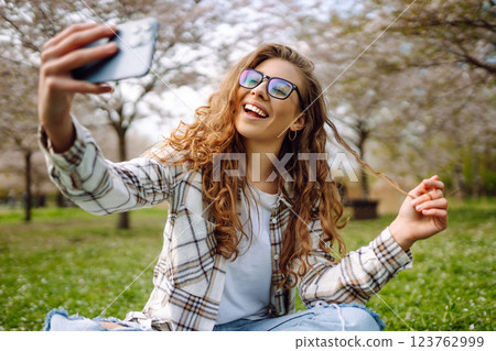 Selfie time. Young woman takes selfie photo in blooming spring park. Concept of relaxation, walking. Selfie time. Young woman takes selfie photo in blooming spring park. Concept of relaxation, walking. 123762999