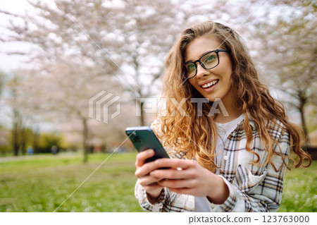 A smiling woman in glasses holds a mobile phone while sitting on a green meadow in a flower park. A smiling woman in glasses holds a mobile phone while sitting on a green meadow in a flower park. 123763000