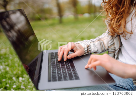 Close-up of woman's hand typing on laptop with notepad at table in park. Concept of remote work. Close-up of woman's hand typing on laptop with notepad at table in park. Concept of remote work. 123763002