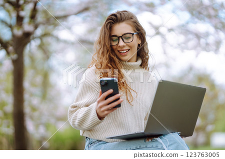 Happy woman with laptop and phone in spring blooming park while working online. Freelancing concept. Happy woman with laptop and phone in spring blooming park while working online. Freelancing concept. 123763005