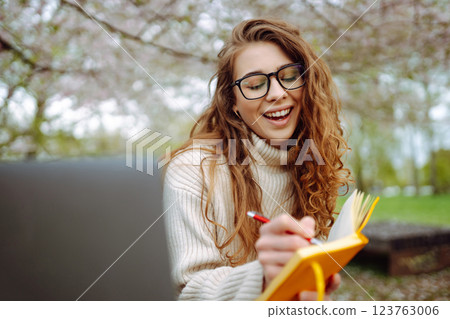 Woman sits on green grass near flowering trees, with laptop, writes in notepad. Planning concept Woman sits on green grass near flowering trees, with laptop, writes in notepad. Planning concept 123763006