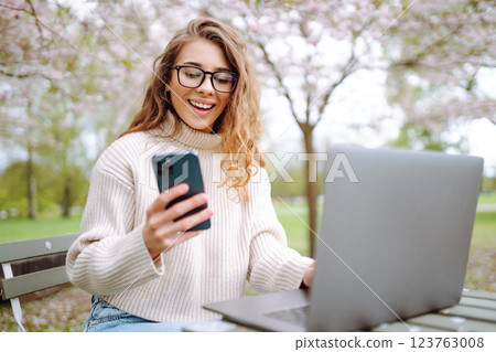 Happy woman with laptop and phone in spring blooming park while working online. Freelancing concept. Happy woman with laptop and phone in spring blooming park while working online. Freelancing concept. 123763008