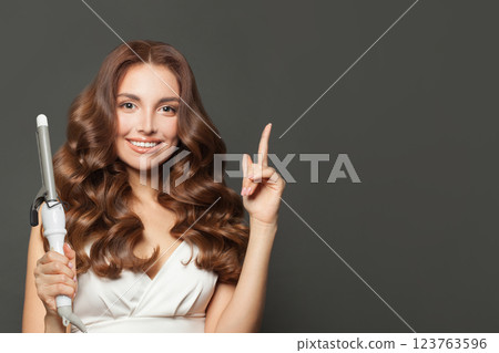 Pointing brunette model using curling iron on grey background.  123763596