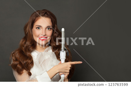 Elegant brown haired woman with shiny long curly hairstyle showing curling iron. 123763599