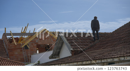 Man standing on roof of house under construction with wooden beams exposed against clear blue sky 123763743