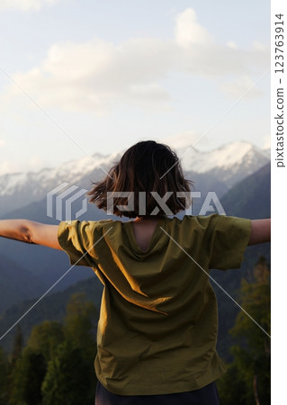 A young woman stands with open arms, enjoying a stunning mountain view. The warm sunlight enhances the scene, symbolizing freedom, adventure, and connection with nature. Travel, mindfulness 123763914