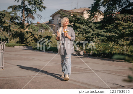 A woman is walking down a sidewalk with a cup in her hand. She is wearing a jacket and jeans. A woman is walking down a sidewalk with a cup in her hand. She is wearing a jacket and jeans. 123763955