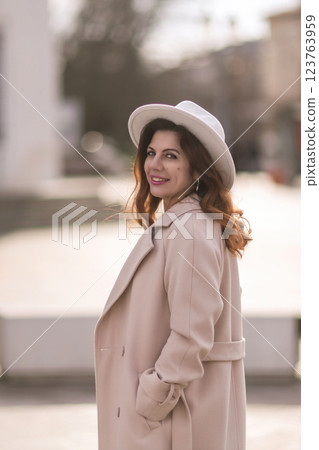 A woman wearing a white hat and a tan coat is standing in front of a building. She has a smile on her face and she is enjoying the moment. 123763959