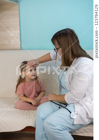 Vertical A female pediatrician examines a sick little girl sitting on a couch at home, touches her forehead to measure her temperature. The pediatrician provides medical care to a sick child at home. 123764129