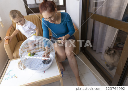 Mother and son feeding a pet budgie in a cage on the balcony Mother and son feeding a pet budgie in a cage on the balcony 123764992
