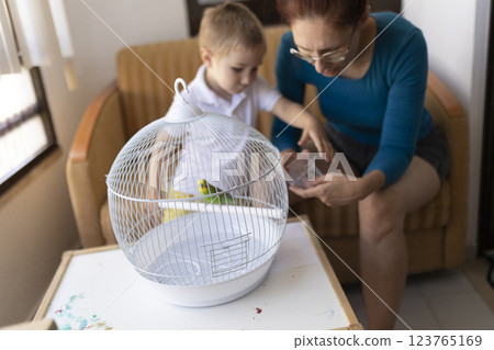 Mother and son caring for pet budgie in white cage 123765169