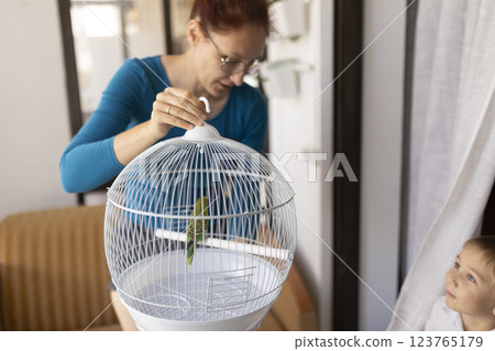 Woman closing birdcage with green parrot inside, child observing Woman closing birdcage with green parrot inside, child observing 123765179
