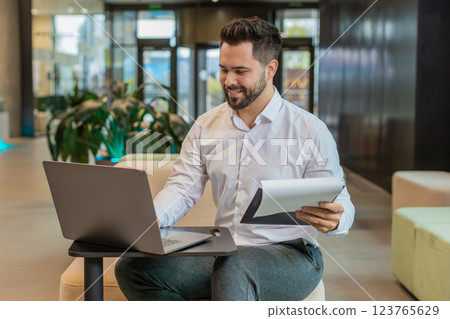 Focused young businessman using laptop and analyzing graph documents reports in modern office lobby Focused young businessman using laptop and analyzing graph documents reports in modern office lobby 123765629