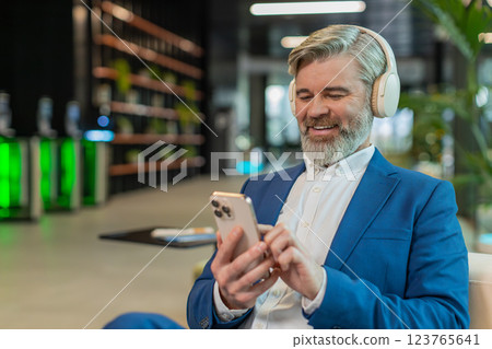 Relaxed middle-aged businessman in headphones listening music on smartphone in modern office lobby Relaxed middle-aged businessman in headphones listening music on smartphone in modern office lobby 123765641